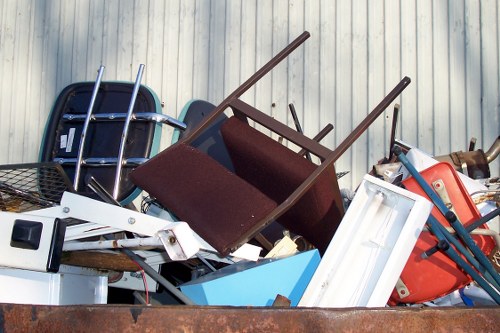 A large pile of discarded household and garden items stacked outdoors against a corrugated metal wall. The collection includes a variety of objects such as a black plastic chair, a brown upholstered armchair with wooden legs, a white window frame, a blue plastic storage container, a white appliance or object, and a red wheelbarrow. The items are arranged in a cluttered manner, with some objects leaning on each other, suggesting they have been collected for rubbish removal. The scene appears to be on a driveway or yard surface, with natural lighting casting soft shadows. This image is representative of waste collection and clearance services that a company like Commercial Waste Abbots Langley might handle in the local area near postcode LU5, illustrating typical household and garden debris waiting to be removed within the Abbots Langley region.