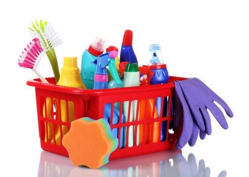 A red plastic basket filled with various cleaning supplies and tools, including multiple spray bottles in blue, red, and green, a yellow bottle, a pink and green scrubbing brush, a blue fabric glove, and an orange sponge with a rainbow-colored edge. The basket is placed on a plain white surface with no background visible, reflecting a clean, organized collection of rubbish and cleaning materials suitable for waste removal services. The assortment of items emphasizes household or commercial cleaning waste, aligned with rubbish disposal and waste management activities often provided by companies like Commercial Waste Abbots Langley. The image's lighting is even, highlighting the vibrant colors and textures of the bottles, brushes, and glove, suggesting a neatly arranged set of cleaning-related rubbish ready for disposal or collection.