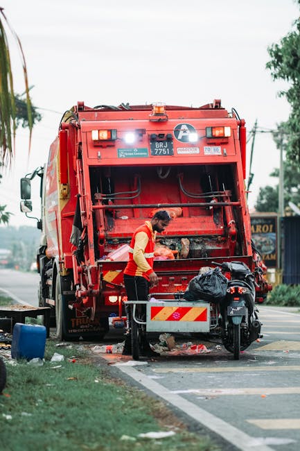 A sanitation worker operating a red garbage truck on a roadside during daytime, with the rear loading mechanism visibly open and engaged in collection. The worker is wearing a high-visibility yellow a