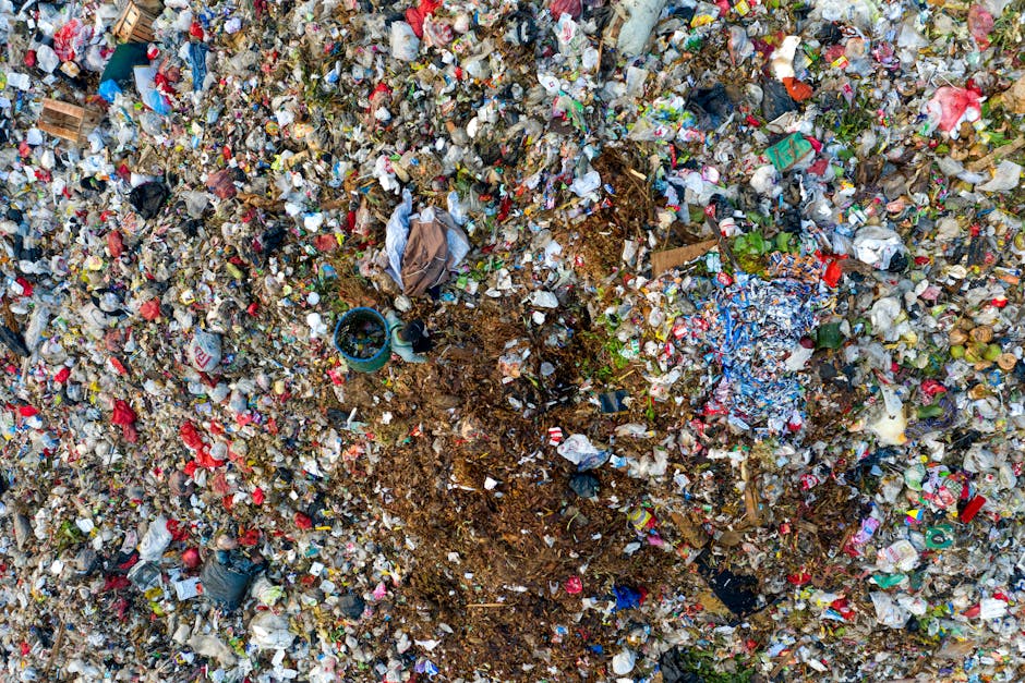 A large landfill site featuring multiple mounds of mixed waste materials, predominantly consisting of compacted gravel and soil covered with assorted colorful plastics, packaging fragments, and discar