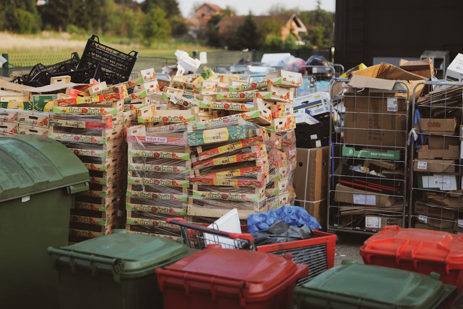 A large outdoor area showing a significant pile of discarded cardboard boxes, primarily from fresh produce packaging, arranged in stacks on a paved surface. The boxes are mostly flat, featuring colorf