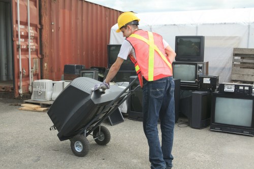 A man wearing a yellow safety helmet, white T-shirt, and high-visibility vest is pushing a black wheeled rubbish bin along an outdoor area with a paved surface. Behind him, there is a large stack of various electronic waste items, including old televisions, monitors, and other appliances, arranged against a red corrugated metal fence. To the right, there are cages containing more waste material, and some wooden pallets are visible nearby. The background features a greenhouse or similar structure made of translucent plastic, suggesting a functional waste collection or disposal site. The overall scene reflects a professional waste management operation related to electronic or general rubbish collection, consistent with services offered by Commercial Waste Abbots Langley in the local area.